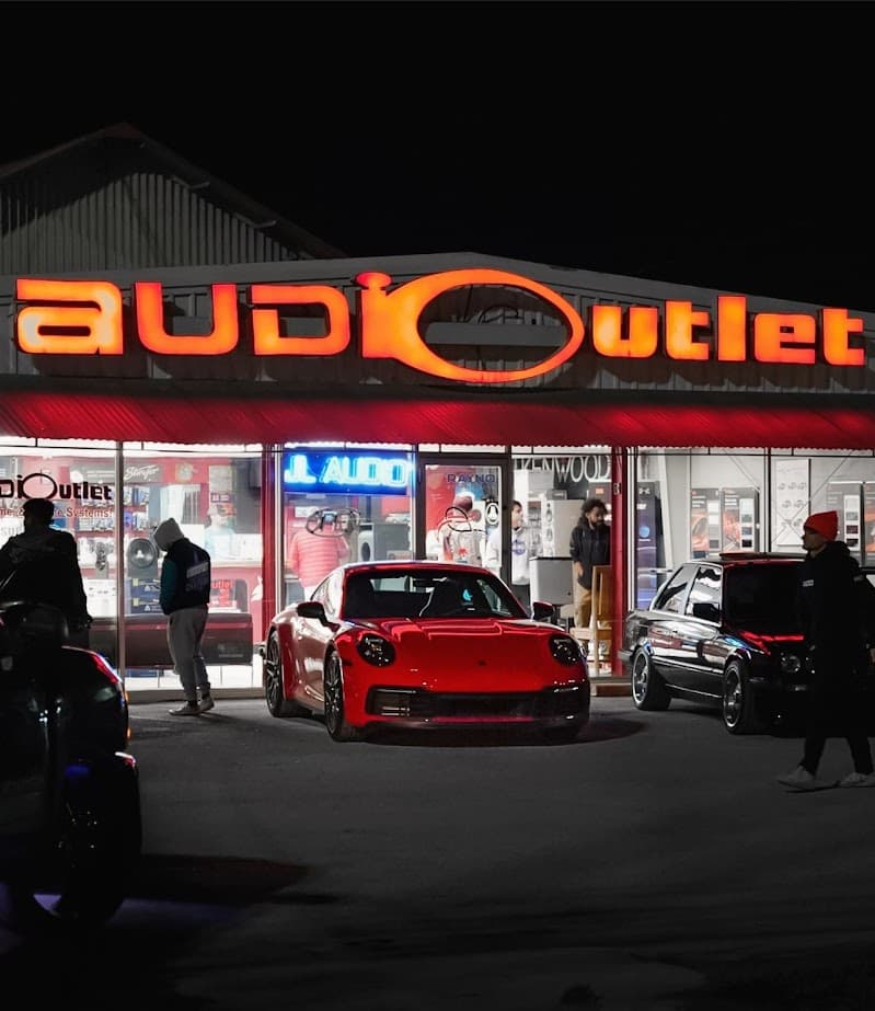 Red sports car parked outside lit storefront after dark at Audio Outlet in San Marcos, Texas