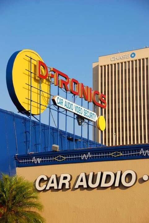 Red and yellow D-Tronics sign above a car audio sign at D-Tronics - Car Audio & Window Tint in McAllen, Texas