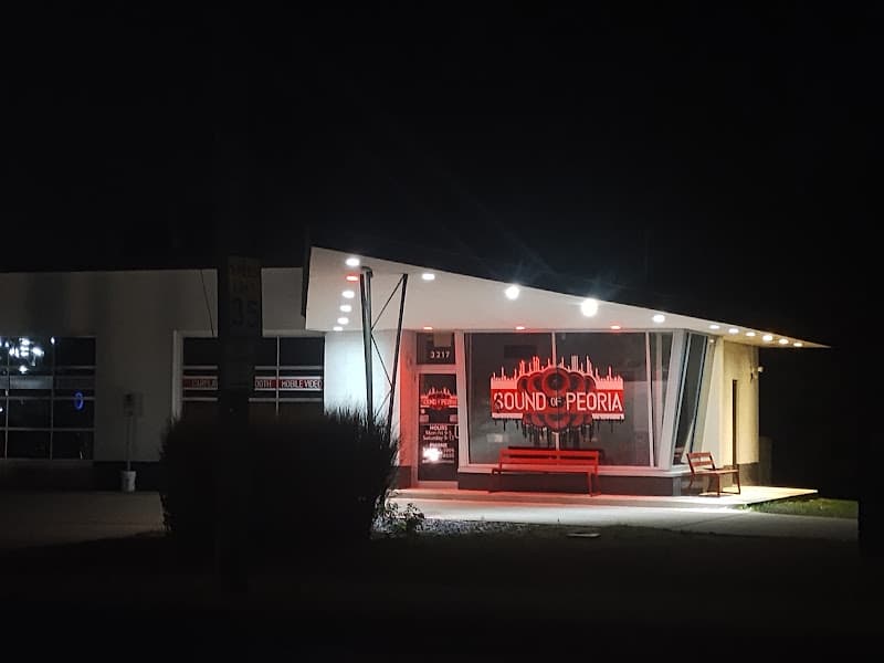 Night exterior of storefront with red neon sign and bench at Sound of Peoria, LLC in Peoria, Illinois