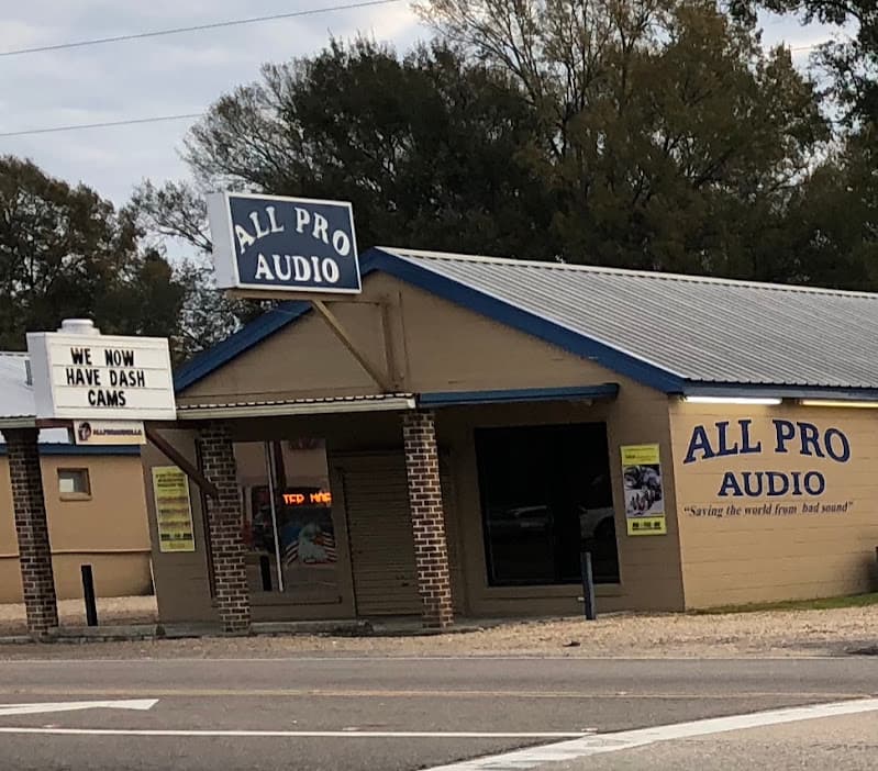 Tan storefront with a roll-up door and blue signs at All Pro Audio in Zachary, Louisiana