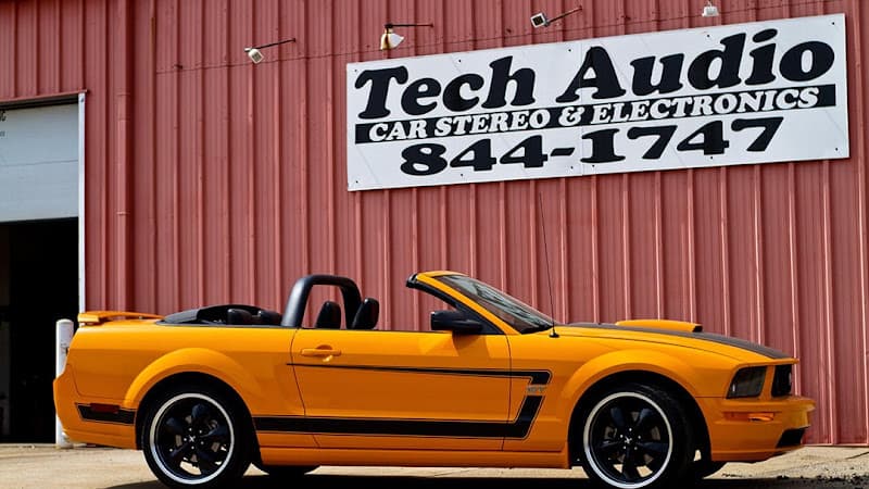 Orange convertible with black stripes parked in front of metal building at Tech Audio in Grand Haven, Michigan
