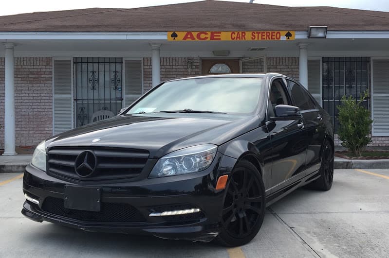 Black Mercedes sedan parked outside a brick shop with a yellow sign at Ace Car Stereo in Houston, Texas
