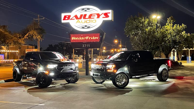 Two lifted black pickup trucks under a lit sign after dark at Baileys Audio and Window Tint in Lake Charles, Louisiana