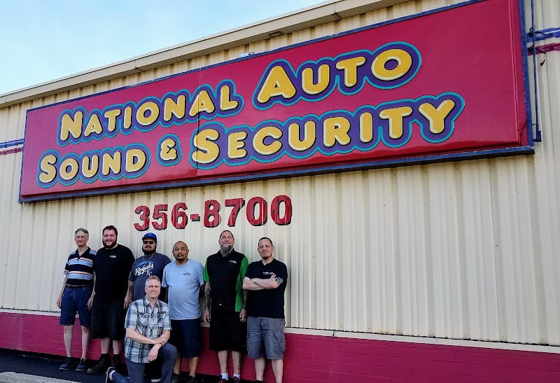 Group of seven men posing in front of a red sign at National Auto Sound & Security in Independence, Missouri
