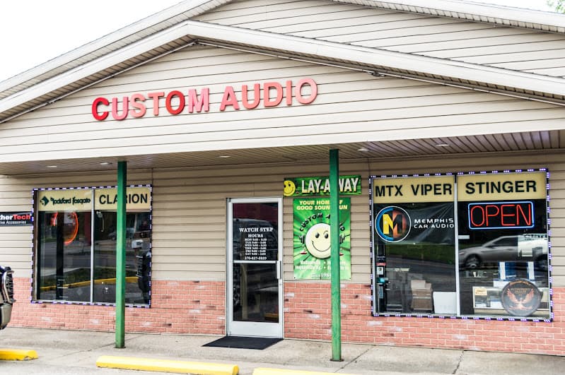 Storefront with red Custom Audio sign and brand decals in windows at Custom Audio in Henderson, Kentucky