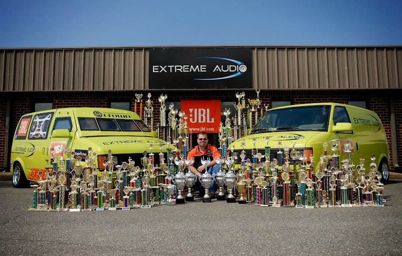 Man kneeling among trophies in front of two yellow vans at Extreme Audio in Mechanicsville, Virginia