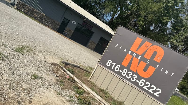 Roadside sign with orange logo on gravel lot at Alarms & Tint KC in Independence, Missouri
