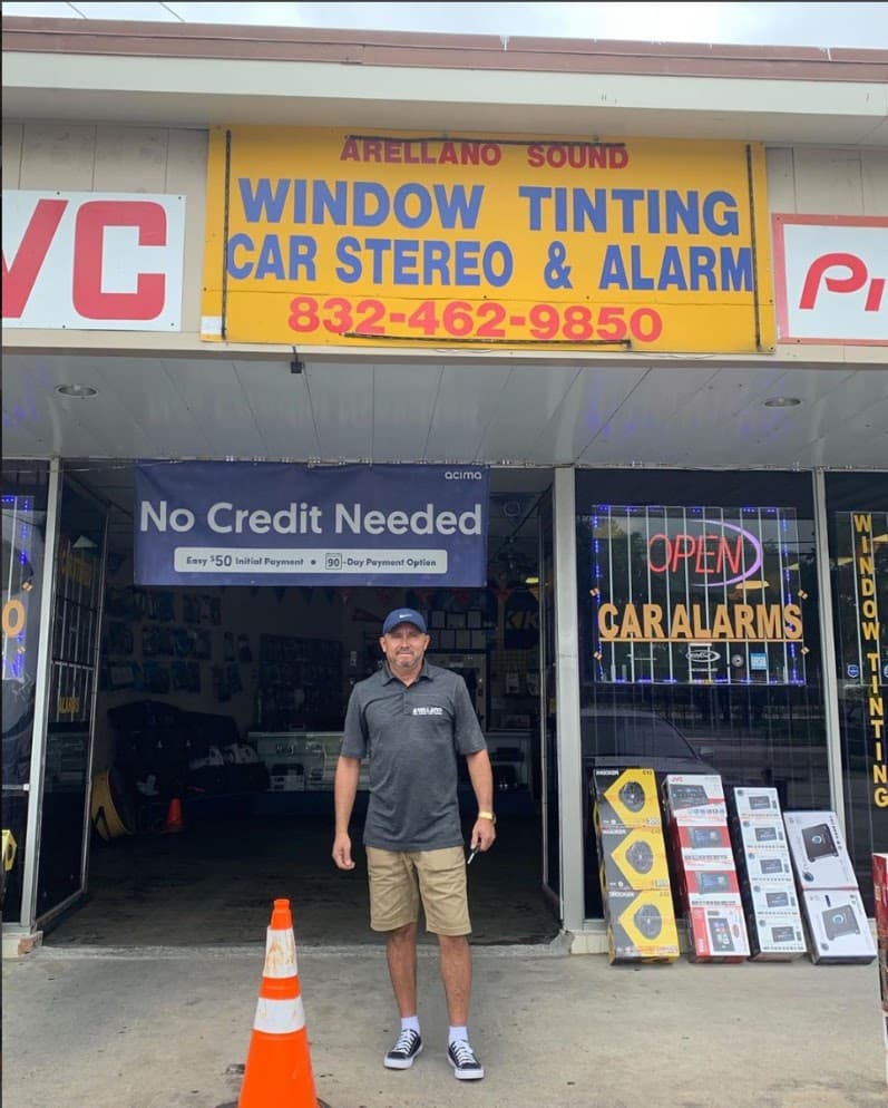 Man in cap standing outside a store with car alarm and stereo signs at Arellano Sound Car Stereo in Houston, Texas